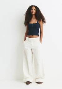 Vrouw met lang krullend haar staat tegen een witte muur, gekleed in een marineblauwe crop top, brede witte broek en bruine sneakers met handen in de zakken.