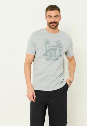 Man with mustache wearing light gray t-shirt with architectural and palm tree design, black pants, standing against plain background.