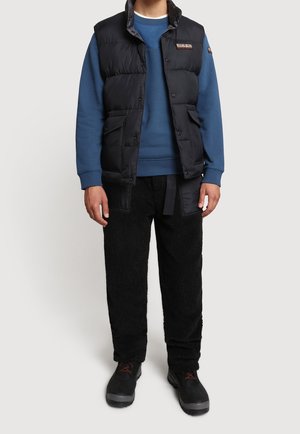 Czarna pikowana kamizelka na niebieskiej bluzie, zestawiona z teksturowanymi czarnymi spodniami. Buty czarne z czerwonymi akcentami. Codzienny, warstwowy strój.