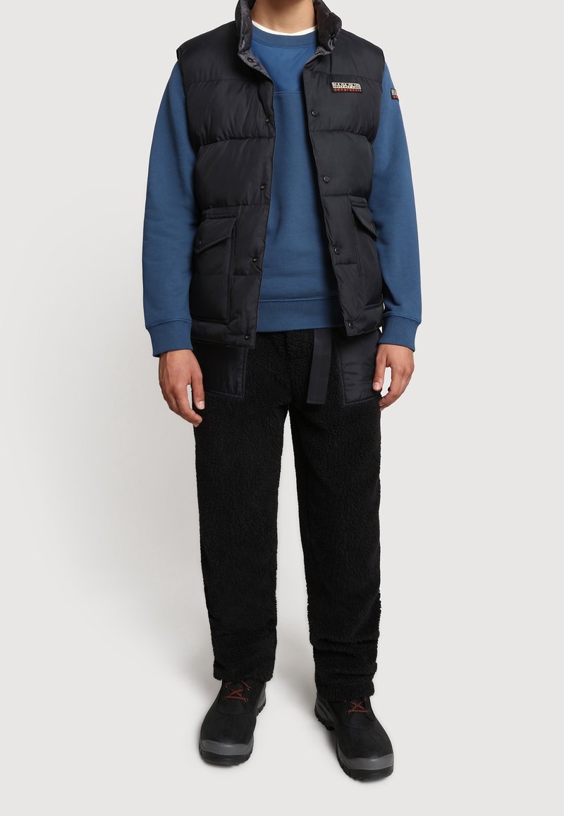 Sort dunvest over blå sweatshirt, kombineret med teksturerede sorte bukser. Skoene er sorte med røde detaljer. Afslappet, lagdelt outfit.