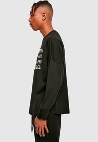 Schwarzes Langarmshirt mit lockerer Passform, das auf der Vorderseite weißen Drucktext aufweist. Rundhalsausschnitt und leicht fallende Schultern.