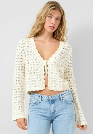 Młoda kobieta o falujących blond włosach, ubrana w kremowy, dziergany sweter zawiązany na piersi oraz jasnoniebieskie, wysokozakładane dżinsy, na jednolitym tle.