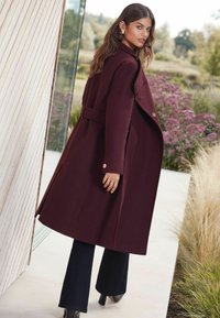 Burgundi gyapjú kabát széles gallérral, övvel a deréknál és aranygombokkal, fekete harangszárú nadrággal és magassarkú cipővel párosítva.