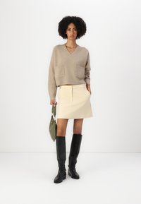Maglione a coste beige con scollo a V e due Tasche frontali, abbinato a una gonna corta color crema e stivali neri alti fino al ginocchio. Borsa verde.