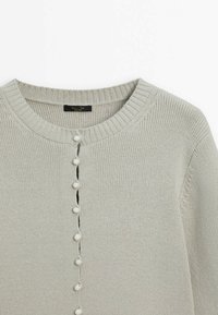 Vaaleanharmaa neulottu villapaita pyöreällä pääntiellä ja etupuolella valkoinen nappirivi. Varustettu resorikäänteillä ja -helmalla.