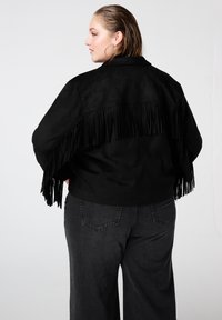 Zwarte suède jas met franjes aan de achterkant, gecombineerd met hooggetailleerde grijze broek. Het ontwerp heeft een losse pasvorm en brede mouwen.