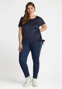 Top corto in satin blu navy con colletto rotondo e dettaglio a fiocco laterale, abbinato a jeans skinny blu scuro con zip laterali dorate e sneakers bianche.