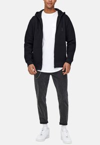 Zwarte zip-up hoodie met een voorzak, gedragen over een wit T-shirt. Gecombineerd met donkergrijze jeans en witte sneakers. Soepele textuur.
