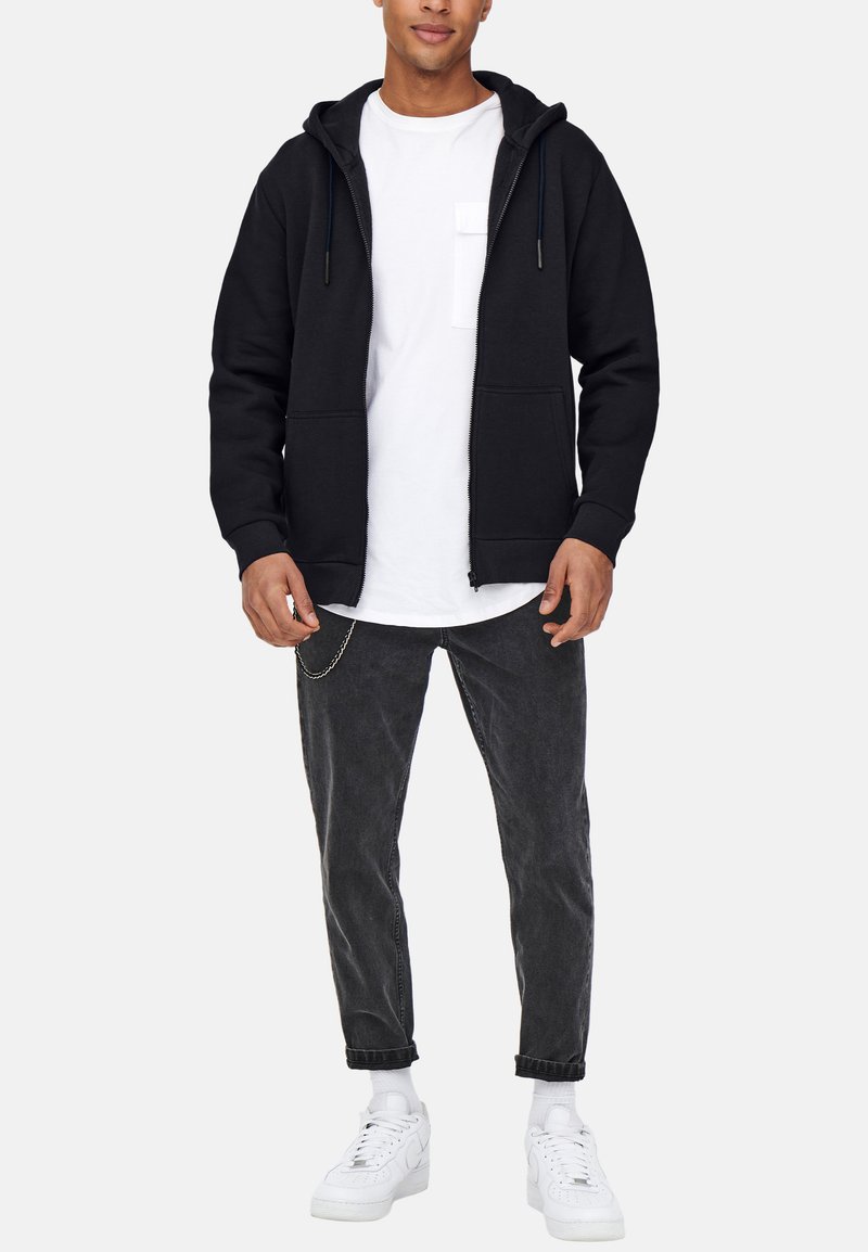 Zwarte zip-up hoodie met een voorzak, gedragen over een wit T-shirt. Gecombineerd met donkergrijze jeans en witte sneakers. Soepele textuur.