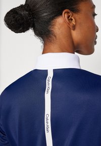 Námořnické polo tričko s bílým límcem a vertikálním bílým pruhem s logem "Calvin Klein". Hladká textura, klasický střih a moderní design.