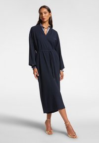 Marineblauwe maxi-jurk met lange mouwen, een tailleband en contrasterende kraagdetails. Soepele stof met een relaxte pasvorm en een enkellange ontwerpe.