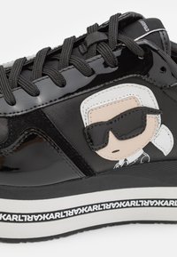 Sneaker in vernice nera con patch di un personaggio dei cartoni animati, lacci neri piatti, suola bianca a contrasto con testo e dettagli testurizzati.