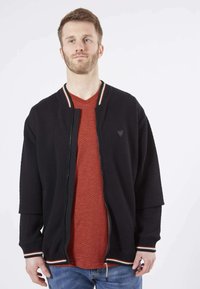Veste noire matelassée avec fermeture éclair à l'avant, col côtelé et poignets rayés, portée sur un t-shirt rouge à col en V, associée à un jean bleu.