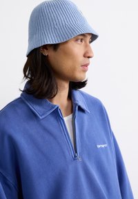 Light blue knitted bucket hat and a blue quarter-zip sweatshirt with a textured finish and embroidered logo on the chest.