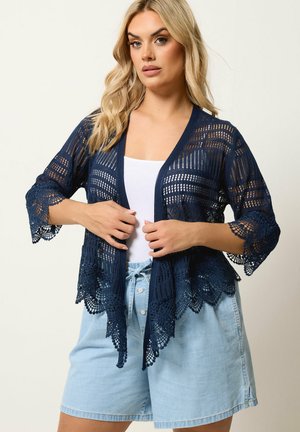 Blonde Frau trägt eine marineblaue Spitzen-Cardigan offen über einem weißen Top und hellblauen hoch taillierten Jeansshorts, die Hände richten die Cardigan.