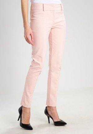 Mujer con pantalones rosa pálido de corte ajustado y zapatos de tacón negro con punta fina, de pie sobre un fondo claro.