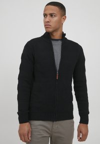 Čierny pletený sveter na zips s textúrovanými horizontálnymi pruhmi, s vysokým golierom a rebrovanými manžetami. Model má na sebe pruhované tričko.