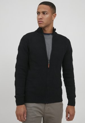 Sort strikket zip-up sweater med teksturerede horisontale striber, med en høj krave og ribbede manchetter. Modellen har en stripet T-shirt på under.