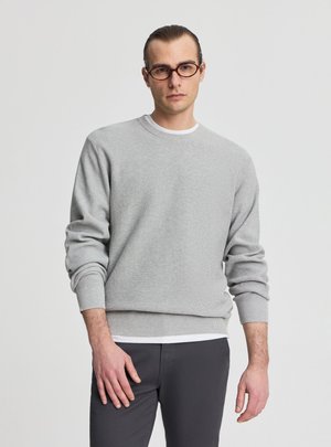 Giovane uomo con capelli pettinati all'indietro, che indossa occhiali tondi, un maglione grigio chiaro sopra una camicia bianca e pantaloni scuri, in piedi davanti a uno sfondo semplice.