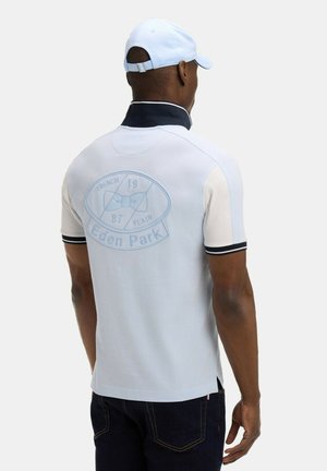 Homme portant un polo bleu clair avec un col bleu marine et le logo « Eden Park » dans le dos, associé à un jean foncé et une casquette blanche, vu de dos.