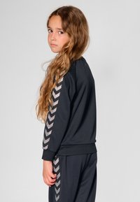 Czarny sportowy strój z akcentami w kształcie jodełki z błyszczącego materiału na rękawach i bokach. Projekt cechuje się luźnym krojem i elastycznymi mankietami.