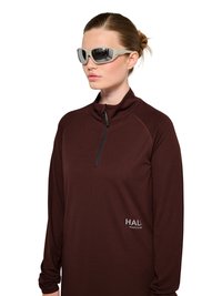 Kvinde med lys hud og knoldet hår bærer beige ombøjede solbriller og en mørkebrun tekstureret trøje med lynlås med teksten "HALA".