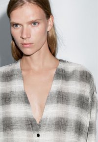 Czarna i biała koszula w kratę z głębokim dekoltem w kształcie litery V, teksturowana tkanina z poziomymi paskami, detal z guzikami z przodu.