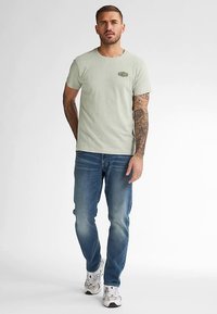 Lichtgroen katoenen t-shirt met klein logo, gecombineerd met blauwe spijkerbroek. De outfit wordt afgemaakt met witte sneakers.