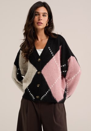 Frau mit langen dunklen Haaren, die eine schwarze, pinke und graue Argyle-Strickjacke mit Knöpfen und dunkle Hose vor einem schlichten Hintergrund trägt.