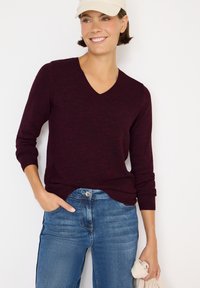 Maglione bordeaux a V con texture a coste e maniche lunghe, abbinato a jeans blu medio. Mani in tasche; tiene un panno di colore chiaro.