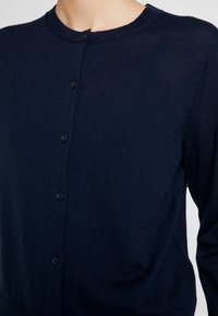 Cardigan blu navy con collo rotondo, texture a coste e chiusura con otto bottoni. Presenta maniche lunghe e tessuto morbido e aderente.