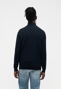 Laivastonsininen neulepaita, jossa on korkea kaulus, sileä rakenne ja vartaloa myötäilevä leikkaus. Yhdistetty vaaleanpesuisiin farkkuihin.