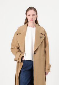 Cappotto lungo in lana di cammello con revers oversize, chiusura a un bottone e tasche laterali; abbinato a un top bianco e jeans scuri.