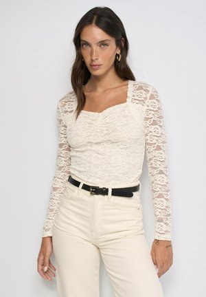Mujer con una blusa blanca de encaje de manga larga con escote cuadrado, pantalones crema de talle alto y cinturón negro sobre un fondo liso.