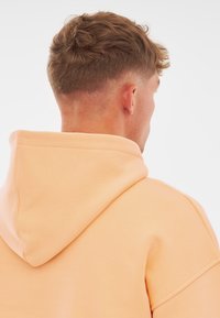 Felpa con cappuccio in morbido tessuto color pesca, caratterizzata da una vestibilità comoda, cuciture a vista e un design semplice. La felpa copre il collo e la schiena.