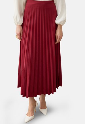 Donna che indossa una gonna midi plissettata rossa a vita alta, una camicetta bianca a maniche lunghe e tacchi alti bianchi con punta appuntita.
