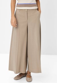 Donna che indossa pantaloni beige a gamba larga e scarpe taupe flat, in piedi con le mani in tasca su uno sfondo bianco.