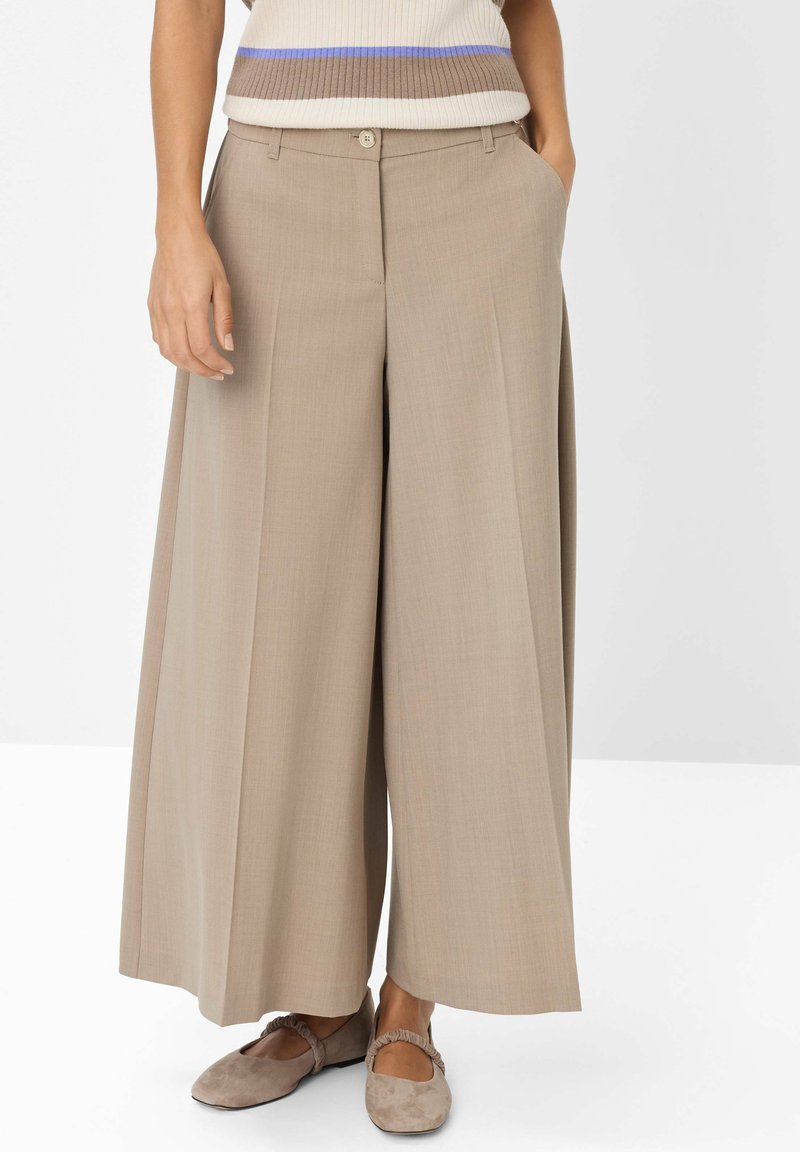 Donna che indossa pantaloni beige a gamba larga e scarpe taupe flat, in piedi con le mani in tasca su uno sfondo bianco.