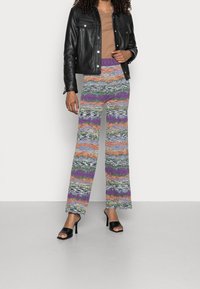 Široké kalhoty s vzorem, multicolor pruhy v purpurové, oranžové, zelené a modré barvě, v kombinaci s černou koženou bundou a černými podpatky.