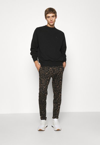Sweatshirt noir à col rond, associé à un pantalon noir ajusté arborant un motif abstrait marron, et des chaussures de sport blanches.