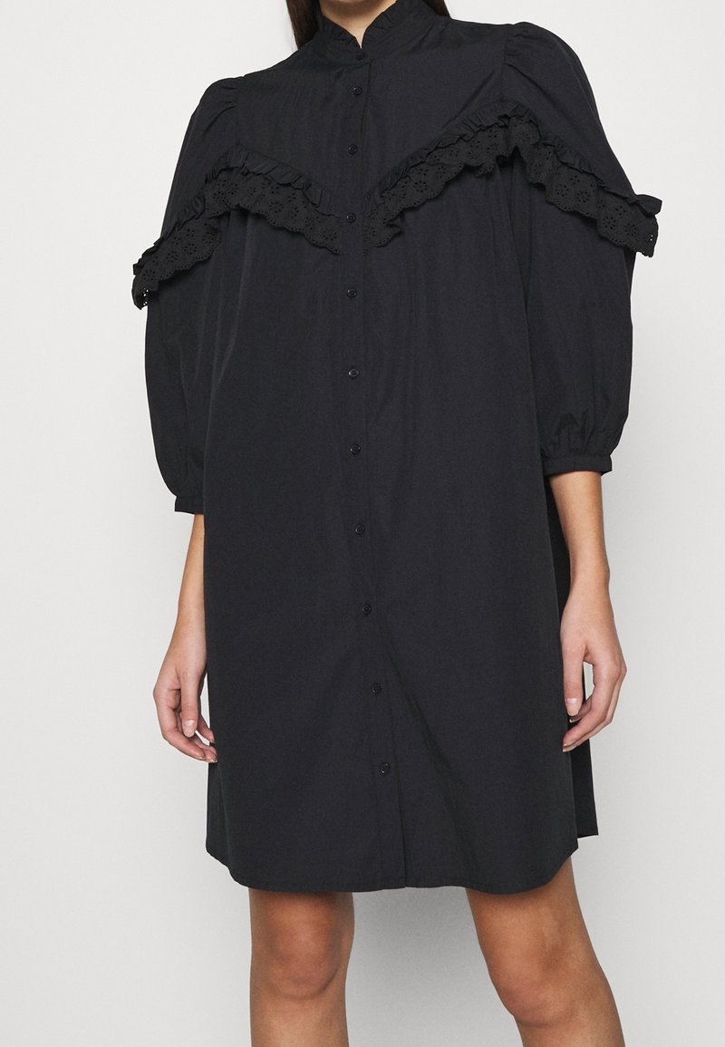 Robe noire boutonnée avec manches trois-quarts bouffantes et volants en dentelle ajourée sur la poitrine et les épaules.