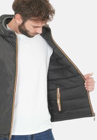Giacca puffer nera con interno grigio, caratterizzata da una chiusura a zip colorata e una tasca laterale con design a strisce. Fodera trapuntata.