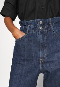 Jeans med rynkad elastisk midja, två sidfickor, mörkblå färg och kontrasterande gula sömmar.