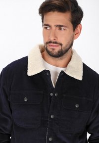 Námorní bleděmodrý kordovým kabát s krémovým umělým beránkem na límci, který má dvě náprsní kapsy a zapínání na patentky. Měkká textura s vertikálními pruhy.