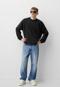 Črna majica s dolgimi rokavi, v kombinaciji s svetlo modrimi širokimi jeans hlačami in belimi supergami. Oseba nosi črne sončne očala.