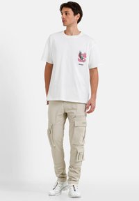 Witte katoenen T-shirt met een grafisch ontwerp op de borst, gecombineerd met beige cargo shorts met meerdere zakken en ritsen. Witte sneakers.