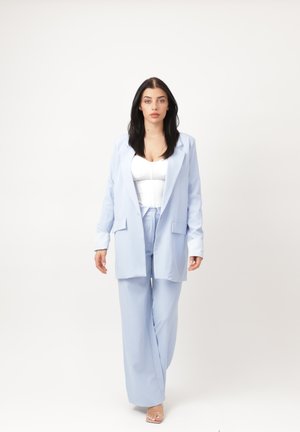 Kobieta z długimi ciemnymi włosami stoi przodem, ubrana w jasnoniebieską marynarkę i spodnie, pod którymi ma biały top, pozując na jednolitym jasnym tle.