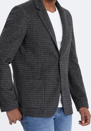 Mężczyzna w ciemnoszarej, kraciastej marynarce na białej koszuli i niebieskich dżinsach, stoi na tle jednolitego białego tła.