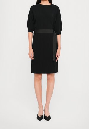 Femme portant une robe noire longueur genou avec des manches courtes bouffantes, une ceinture noire à la taille et des escarpins noirs à bouts pointus.
