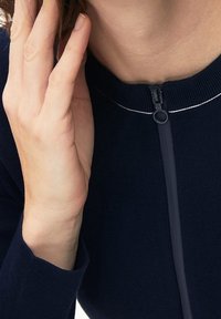 Mörkblå zip-up-tröja med en smidig stickning, kort hals och en silverdetalj längs dragkedjan. Handen vilar nära halsringningen.